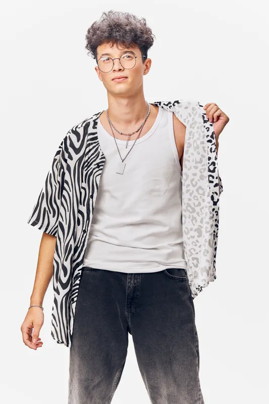 young man in animal print shirt and white tank top wearing glasses with curly hair, posing confidently against white background, casual style portrait
