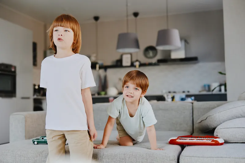 Two boys playing in a bright living room, one standing while the other crawls on the sofa, showcasing playful interaction and a warm home atmosphere.