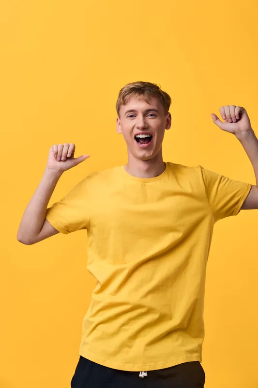 Cheerful man in a yellow shirt raises his arms in celebration against a bright yellow background, radiating energy and positive mood.