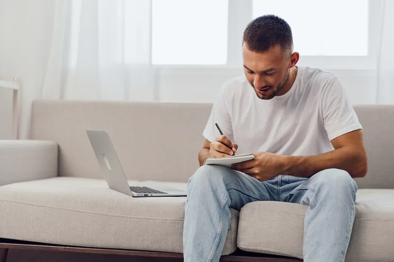 Young man writing in a notebook during a digital detox, reflecting on work-life blur and mental health, embracing authentic imperfection at home.