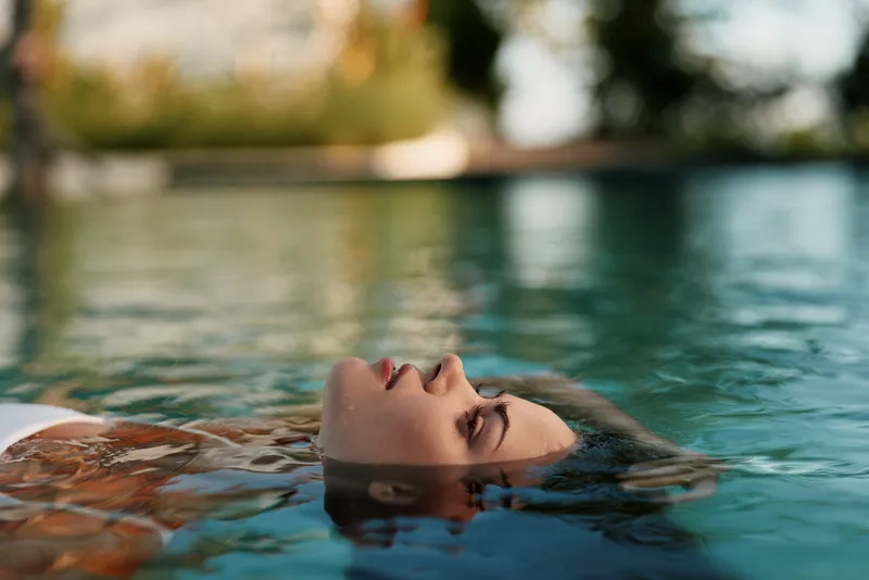 Relaxed person floating on their back in a tranquil pool, water surrounding the face, warm sunlight, calm mood, lifestyle and wellness vibe, subtle self care atmosphere today