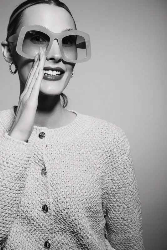 Young white woman with fair skin wearing oversized sunglasses smiles and gestures near mouth in studio. Concept of fashion, style, and happiness.