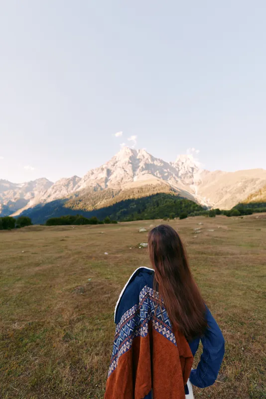 Woman with long hair seen from back wearing a patterned shawl stands in a grassy meadow facing a towering mountain landscape, travel adventure, solitude and wide open nature view.