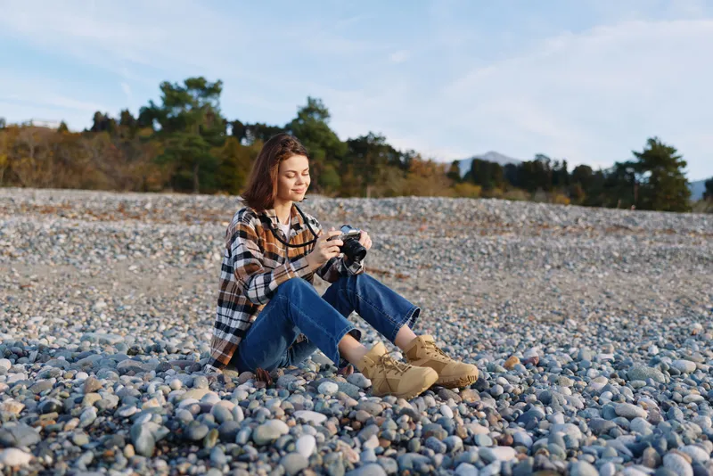 photographer, camera, beach, outdoor, nature, woman sitting on pebbled shore adjusting gear for a candid shot, casual plaid shirt, jeans and boots under soft autumn light, calm coastal scene