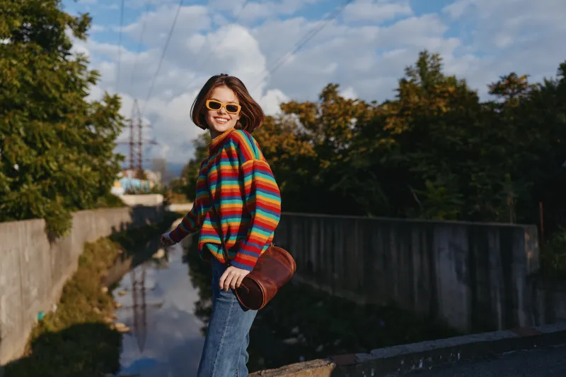 woman in a colorful striped sweater and sunglasses smiling by a canal in an urban setting, wearing jeans and a crossbody bag, leaning on a railing under blue sky, lifestyle travel vibe
