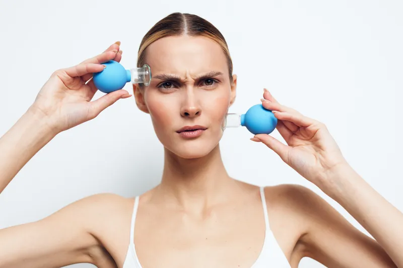 Young woman with blonde hair uses blue facial suction cups on her forehead and cheek while frowning against a white background in a skincare routine.