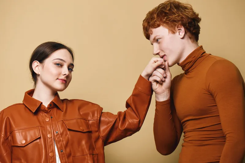 Man and woman with red hair and brown clothes posing together on beige background. Couple showing affection and stylish fashion with confident expressions and close connection.