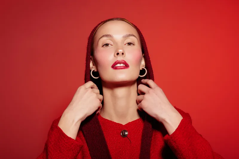Young woman in red sweater and headscarf poses confidently against red background. She wears gold hoop earrings and bright red lipstick. Concept of fashion, beauty, and style.