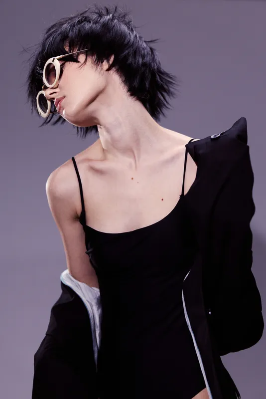 Stylish woman wearing black outfit and oversized sunglasses posing against purple background with edgy hairstyle and confident expression