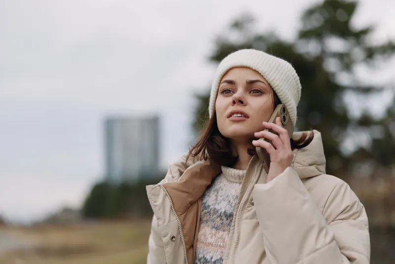 Woman in a warm coat and beanie speaks on the phone outdoors, posing in a misty landscape with trees in the background. Casual chic style and natural light create a calm portrait.