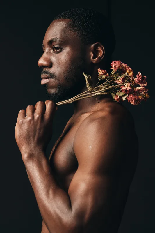Black man with dark skin holding dried flowers on shoulder in creative visual design style, portrait with bare torso and thoughtful expression on black background.