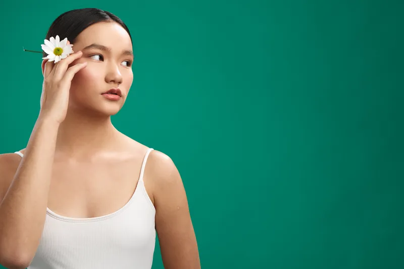 asian woman with a flower tucked behind her ear stands against a bold green backdrop, calm and elegant, creating a serene portrait that highlights natural beauty and thoughtful presence.