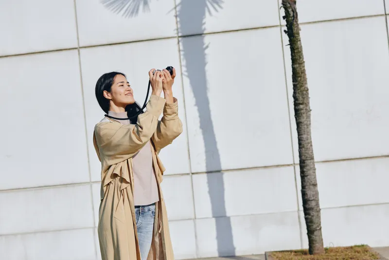 Woman in beige trench coat photographs palm trees against a white wall in an outdoor scene, capturing a candid moment with daylight and urban travel vibes.
