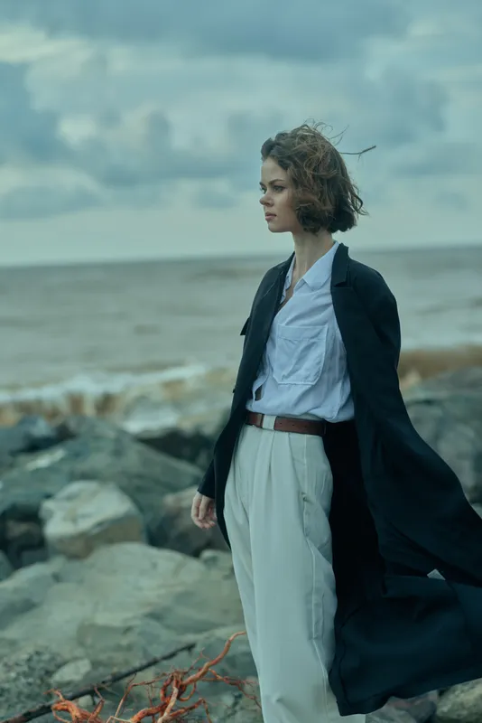 Windy seaside scene with a businesslike woman walking along rocky shore, wearing a long coat, white shirt and belt, ocean backdrop, moody sky, confident stride, fashion meets nature