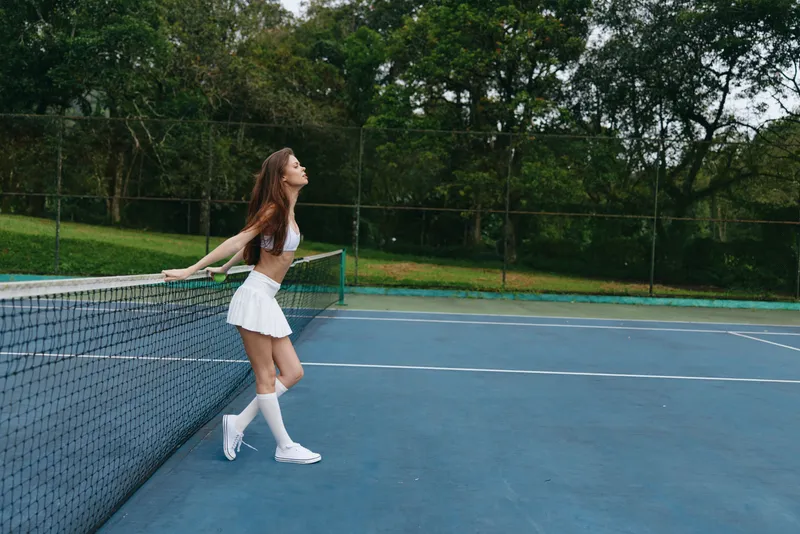 Young female tennis player in white sportswear enjoying a moment on the court, exuding confidence and joy in a serene outdoor setting, perfect for sports themed content