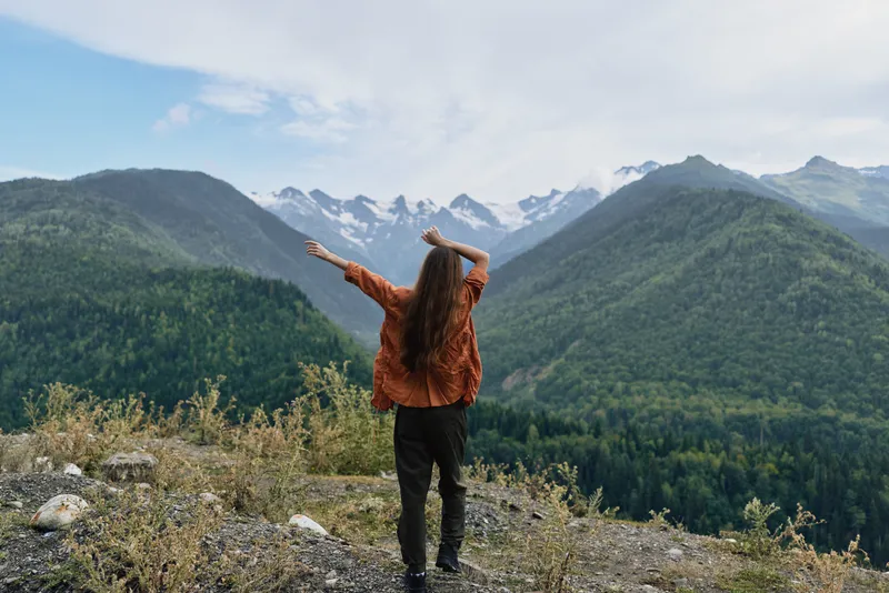 Person standing on a rocky overlook, facing towering alpine mountains with arms raised, capturing a moment of freedom and connection with nature and calm.