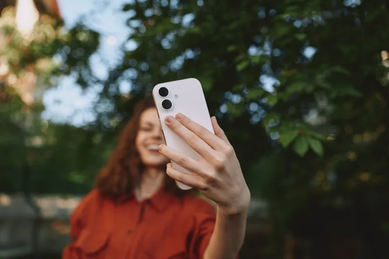 Young woman in an orange shirt takes a candid outdoor selfie, capturing a natural lifestyle moment with a bright smile and a blurred green background of trees