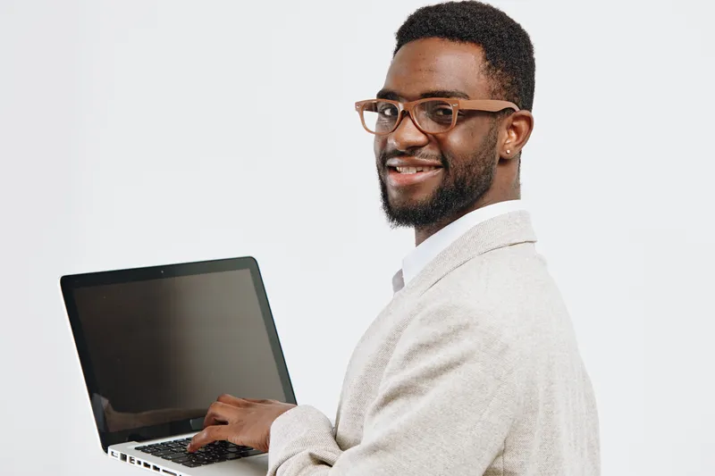 Smiling man with glasses wearing beige blazer holding laptop, looking at camera with confident expression, isolated on plain white background, studio portrait, professional, modern work environment.