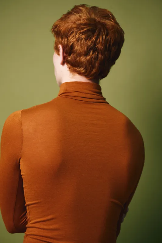 Redhead person with curly hair, wearing orange turtleneck, standing with back to camera against green background. Studio portrait, casual outfit and relaxed posture with natural light.