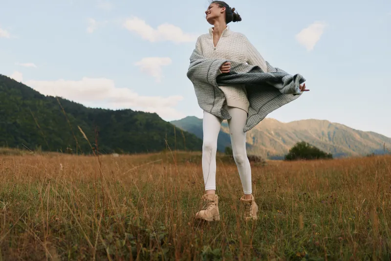 Woman in a light sweater and coat stands in a grassy field near mountains, enjoying fresh air and outdoor lifestyle amid nature.