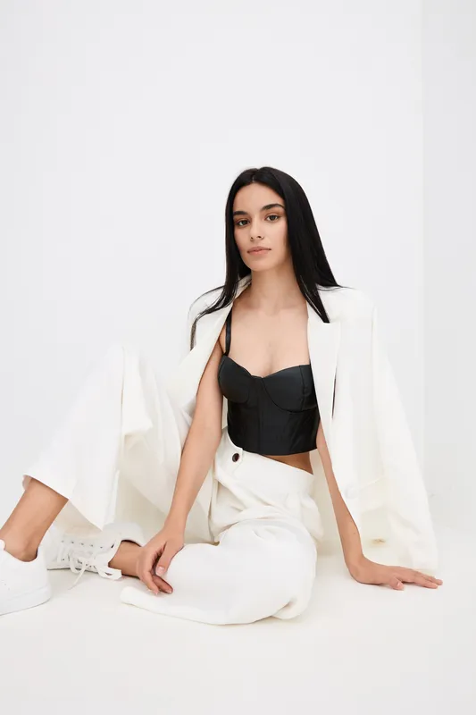 Young woman with long black hair wearing white wide-leg pants, white blazer and black corset top, sitting on white floor, minimalist fashion style, stylish outfit.
