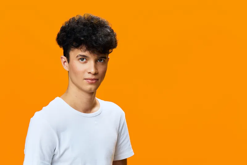 young man with curly hair wearing white t-shirt on bright orange background looking confidently, studio portrait, casual style, isolated vibrant backdrop