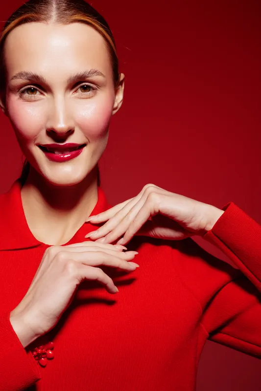 Smiling woman with red lipstick and red sweater poses with hands near face on red background. Concept of beauty, fashion, and confident female portrait with vibrant colors and studio lighting.