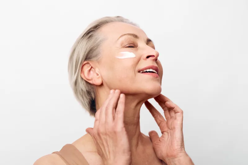 Mature woman with short gray hair applying cream under eyes on clear skin, smiling gently against white background. Concept for skincare, anti aging, wellness, and self care routine.