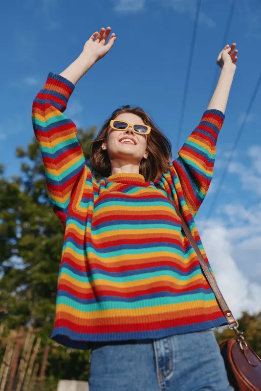 Young woman in a rainbow sweater smiling outdoors with sunglasses and arms raised, casual outfit and denim jeans under blue sky, joyful lifestyle portrait for fashion and travel.