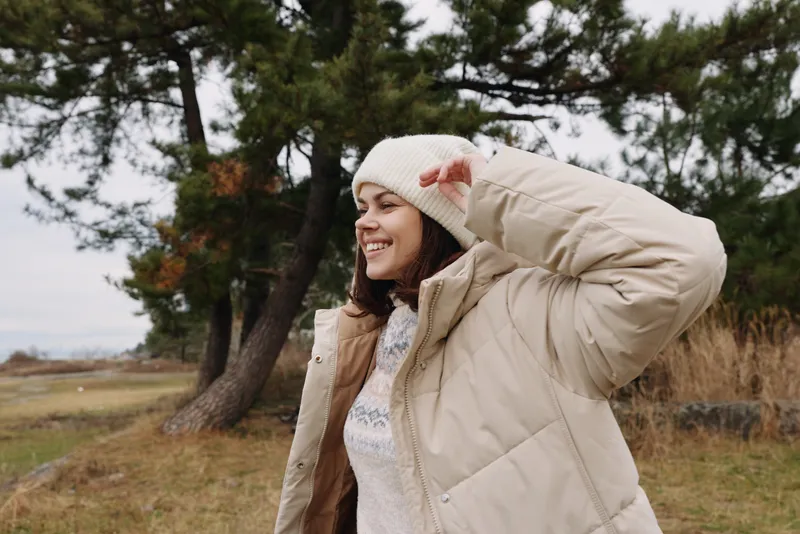 Woman in a warm coat and knit hat enjoying an outdoor scene, standing in a field with trees, smiling softly as she shields her eyes from the breeze.