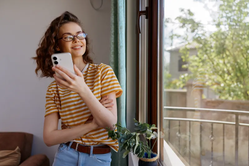 A woman in a striped tee stands by a sunlit window, smiling at her phone, captured in a cozy home lifestyle scene. Casual, everyday moment with digital connection.