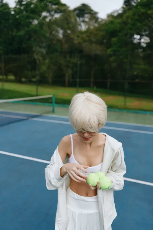 Young woman in white sportswear with short blonde hair, holding green tennis balls on a court, exuding focus and determination during summer sports practice