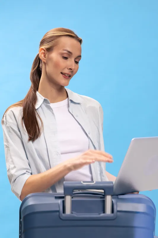 Young woman with long hair stands behind suitcase using laptop. Casual outfit with white shirt and light shirt. Concept for travel planning, business trip, and digital nomad lifestyle.