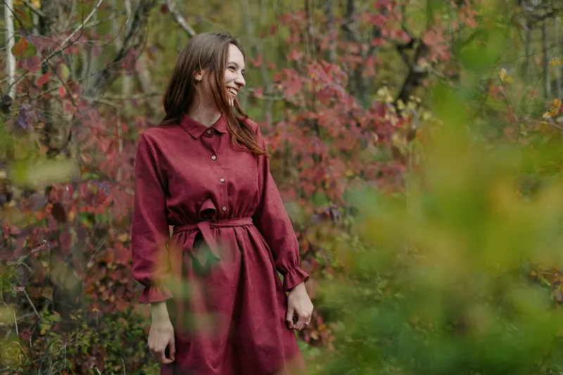 Young woman in a simple red dress stands among autumn foliage, smiling in a natural candid pose that conveys authenticity and credible emotional presence in portraiture.