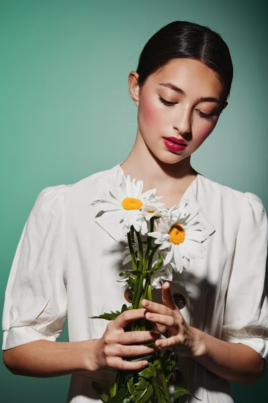 Young woman wears white blouse holding three white daisies, her lips vivid red and eyes gently closed against green background. Concept of beauty fashion and creative portrait.