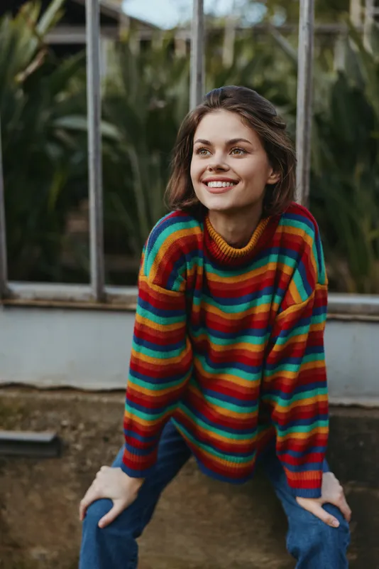 Young adult woman smiling in a rainbow striped sweater and casual outfit, sitting and leaning outdoors by green plants with brown hair, relaxed lifestyle portrait for fashion and travel mood.