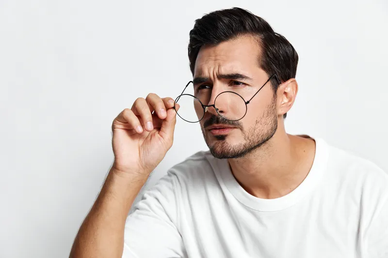 Thoughtful man holding glasses look at camera wearing white t-shirt with serious expression isolated on plain light background. People lifestyle concept.