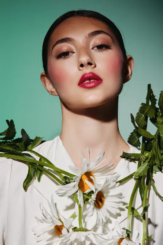 Portrait of a young woman with glossy red lips and bold pink blush wearing a white outfit adorned with daisies on a pale green background. Fashion and beauty creative concept.