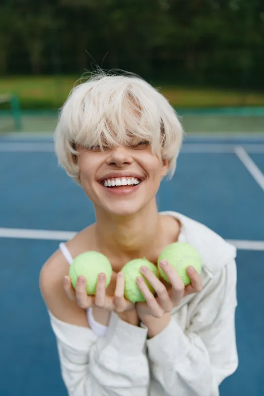 Young woman with short blond hair smiles while holding tennis balls, dressed in casual sporty attire, set on a blue tennis court backdrop