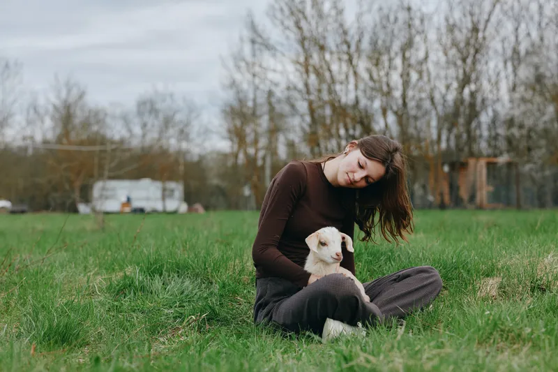 Calm woman sits on grass in a field, cuddling a fluffy white dog in a sunny outdoor scene, expressing gentle companionship and relaxed mood during a casual lifestyle moment.