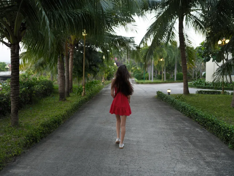 Woman walking alone in a red dress along a palm tree-lined path during summer evening with lush greenery and tranquil atmosphere.