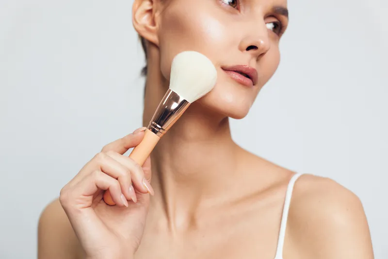 Woman applying makeup with brush holding powder, beauty skincare routine closeup in studio, calm focused expression, smooth skin texture, white background, soft diffused light, natural neutral palette