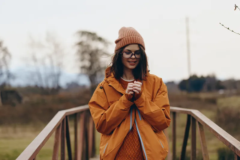 Smiling woman lifestyle outdoors on a wooden bridge, wearing an orange jacket and knit beanie with glasses, autumn scenery and soft background blur, casual travel mood