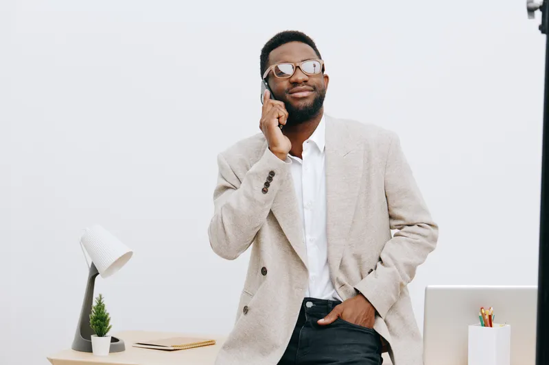 Professional young man wearing glasses talking on smartphone in office environment with white background, smiling confident posture, modern workspace, business casual attire, lifestyle concept