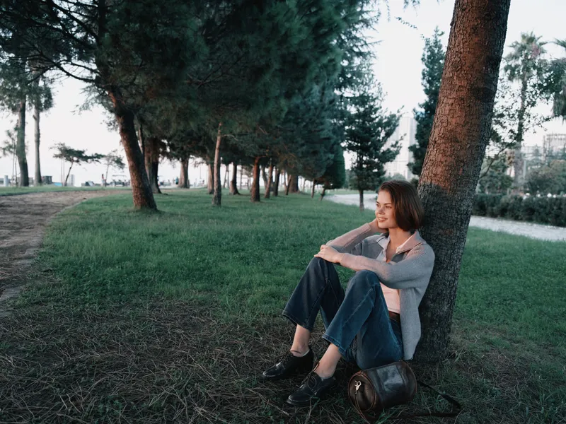 A thoughtful woman sits against a tree in a tranquil park, enjoying a peaceful moment outdoors. She wears casual attire, smiling softly, radiating calm and contemplation.
