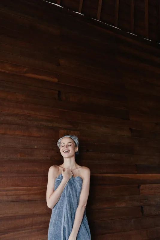 young woman wrapped in towel and hair wrapped against dark wooden wall, relaxed and happy expression, natural light, cozy bathroom atmosphere