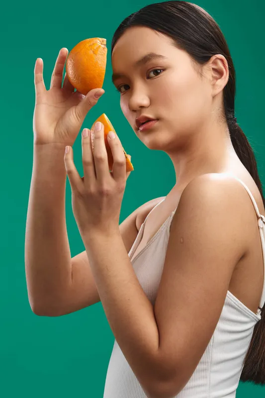 Asian woman holds bright orange slices against a vibrant green backdrop, looking at the camera with a soft smile, presenting a fresh, healthy lifestyle and bold color contrast.