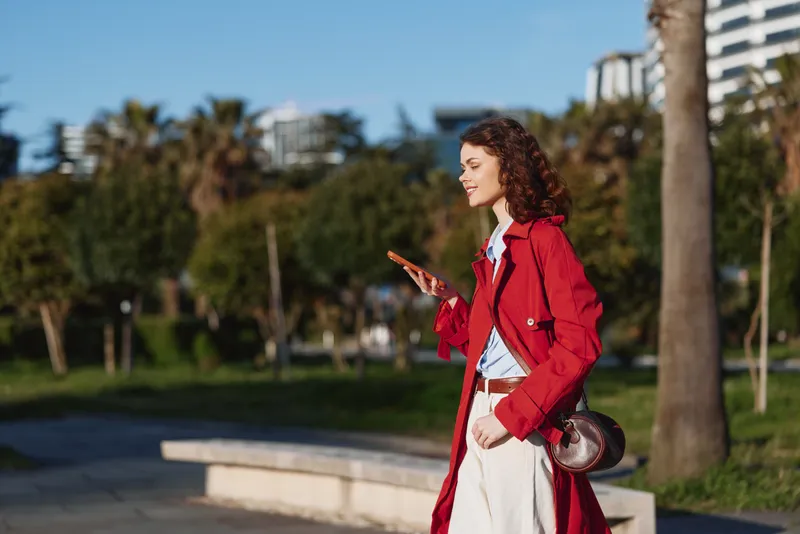 A young woman in a red coat walks outdoors using a smartphone on a sunny day. She smiles while standing near palm trees in an urban park environment.