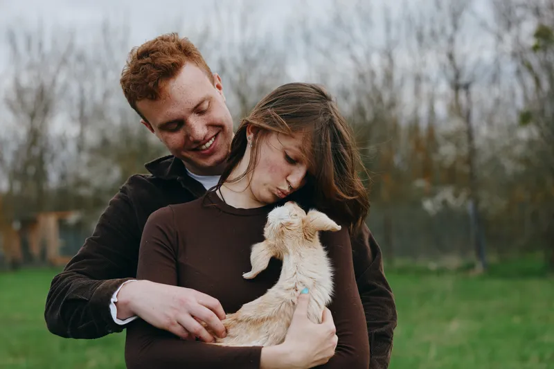 Loving couple outdoors gently holds a small goat kid, showcasing warm affection and rural lifestyle, serene park setting, cozy sweaters, and a peaceful bonding moment in a grassy field.