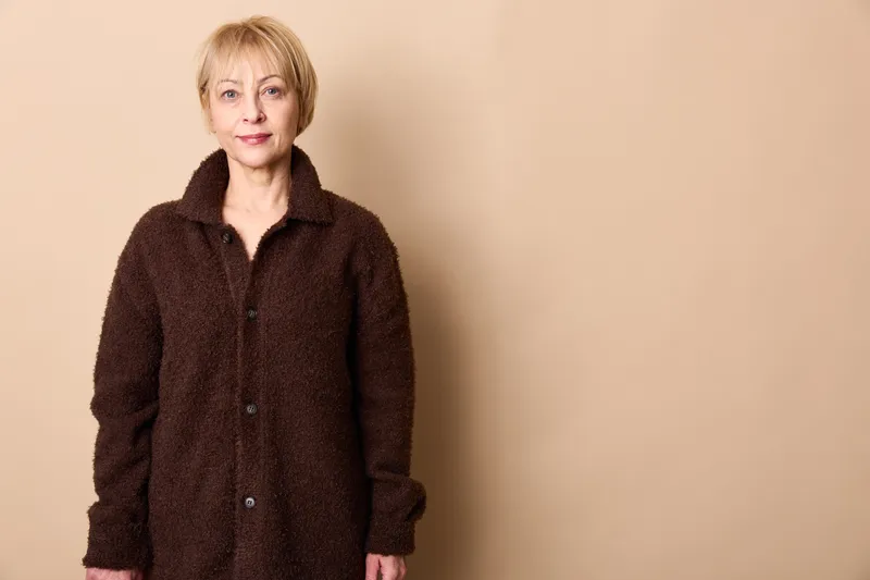 Woman senior elderly portrait standing in a studio, beige background, wearing a dark coat, calm neutral expression, short hair, relaxed posture, copy space for text, dignified calmness